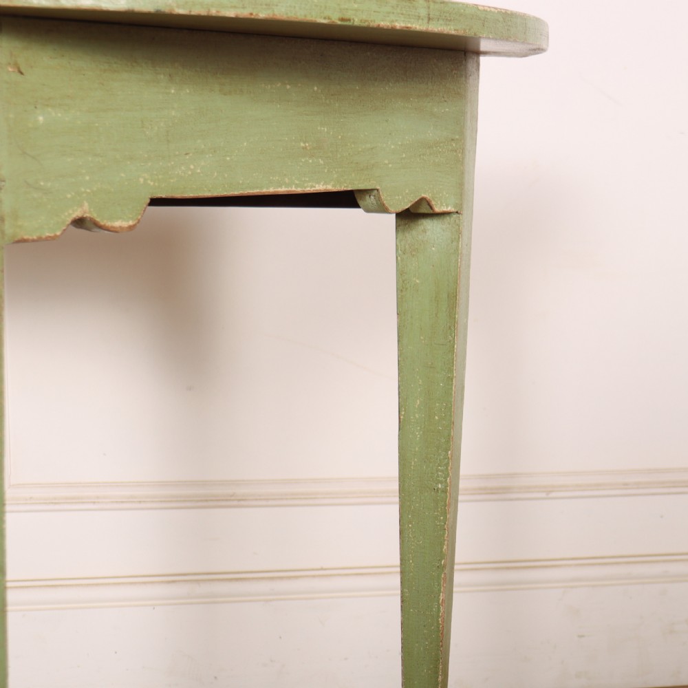 A semi-circular green painted wooden console table featuring three tapered legs and a scalloped apron against a white wall. View II