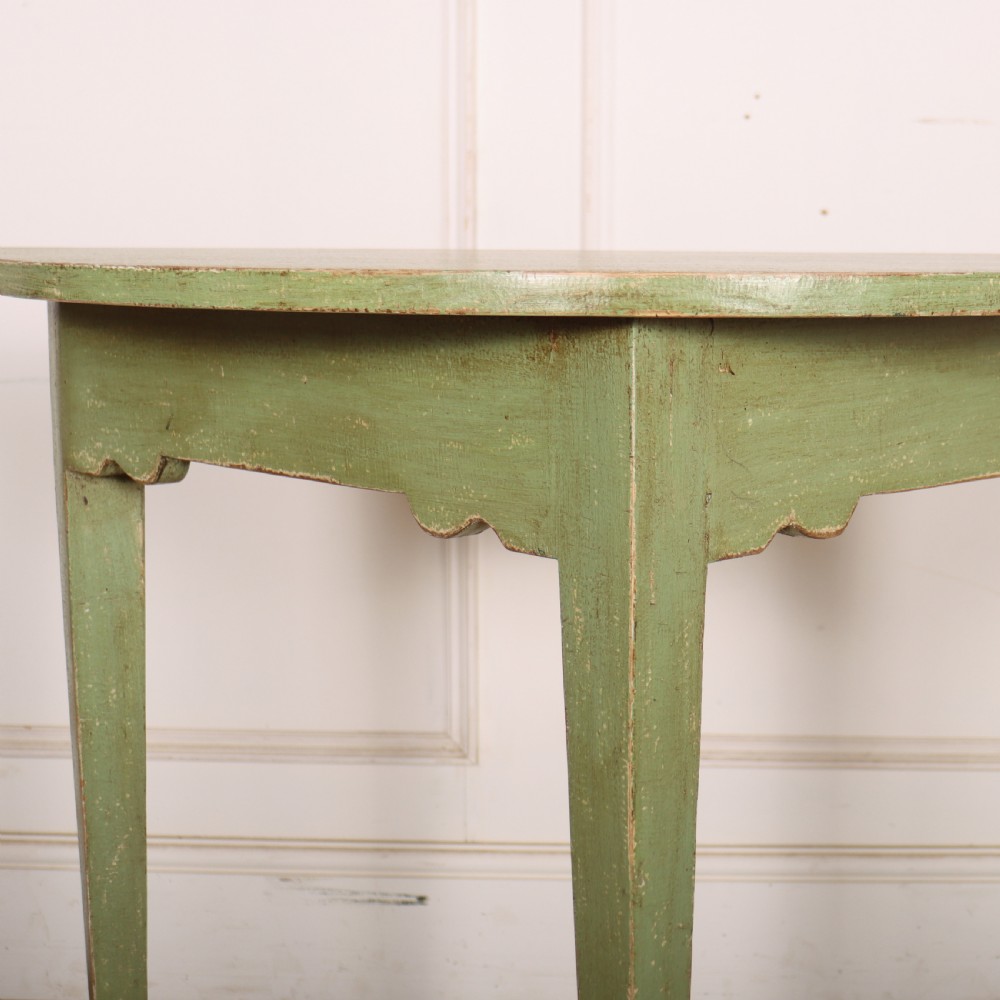 A semi-circular green painted wooden console table featuring three tapered legs and a scalloped apron against a white wall. View III