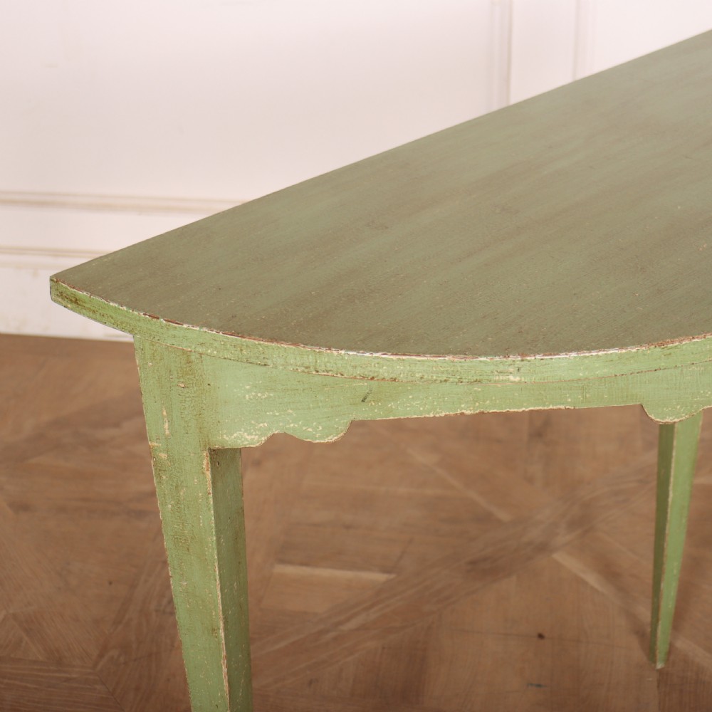 A semi-circular green painted wooden console table featuring three tapered legs and a scalloped apron against a white wall. View V