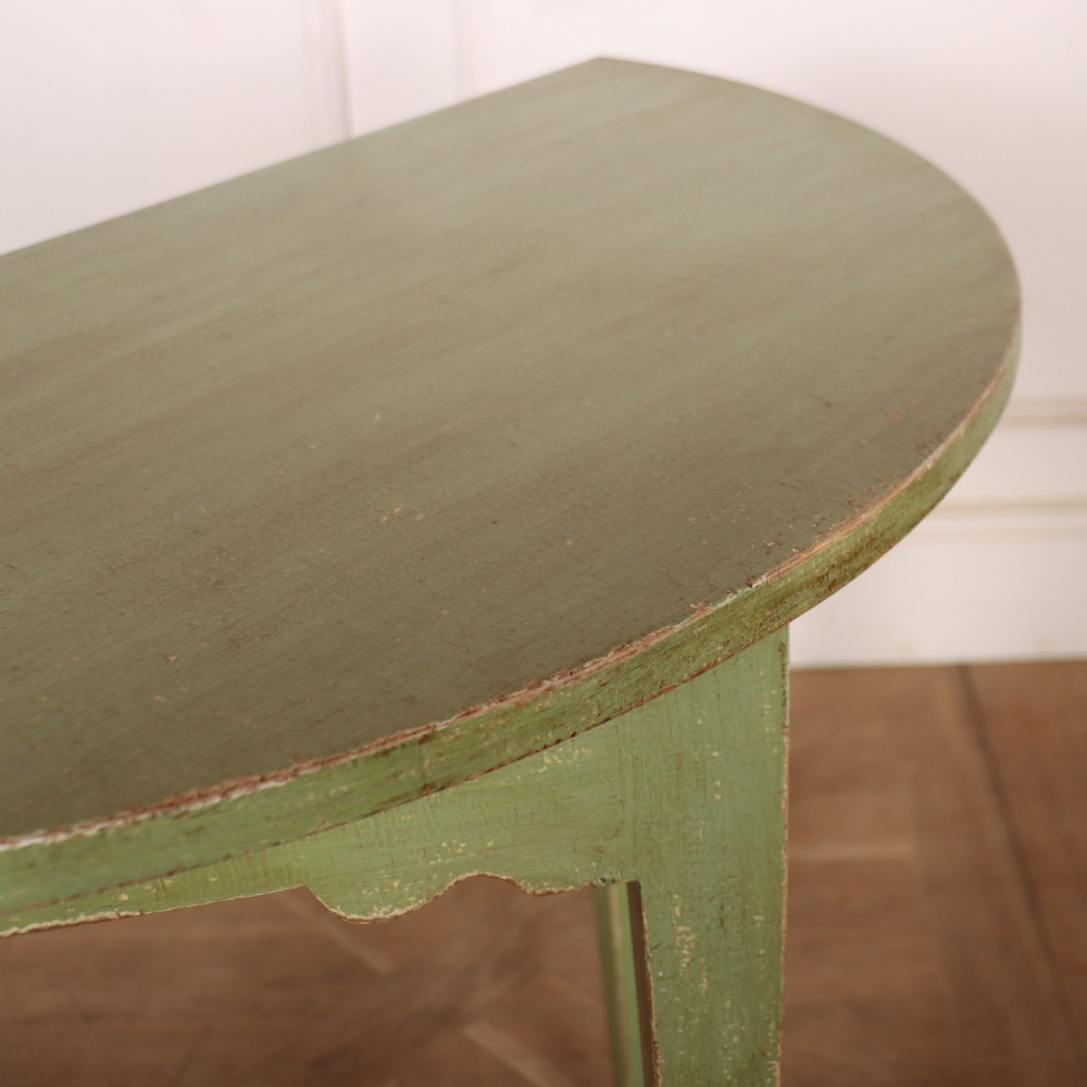 A semi-circular green painted wooden console table featuring three tapered legs and a scalloped apron against a white wall. View VI