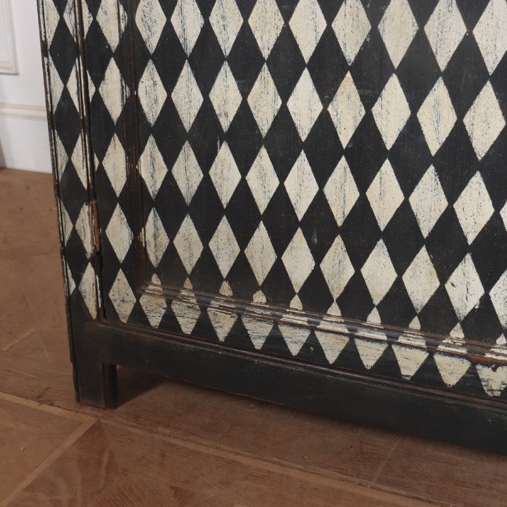 A tall English painted wooden linen cupboard featuring a black and cream harlequin diamond pattern across the front doors and drawers. View XV