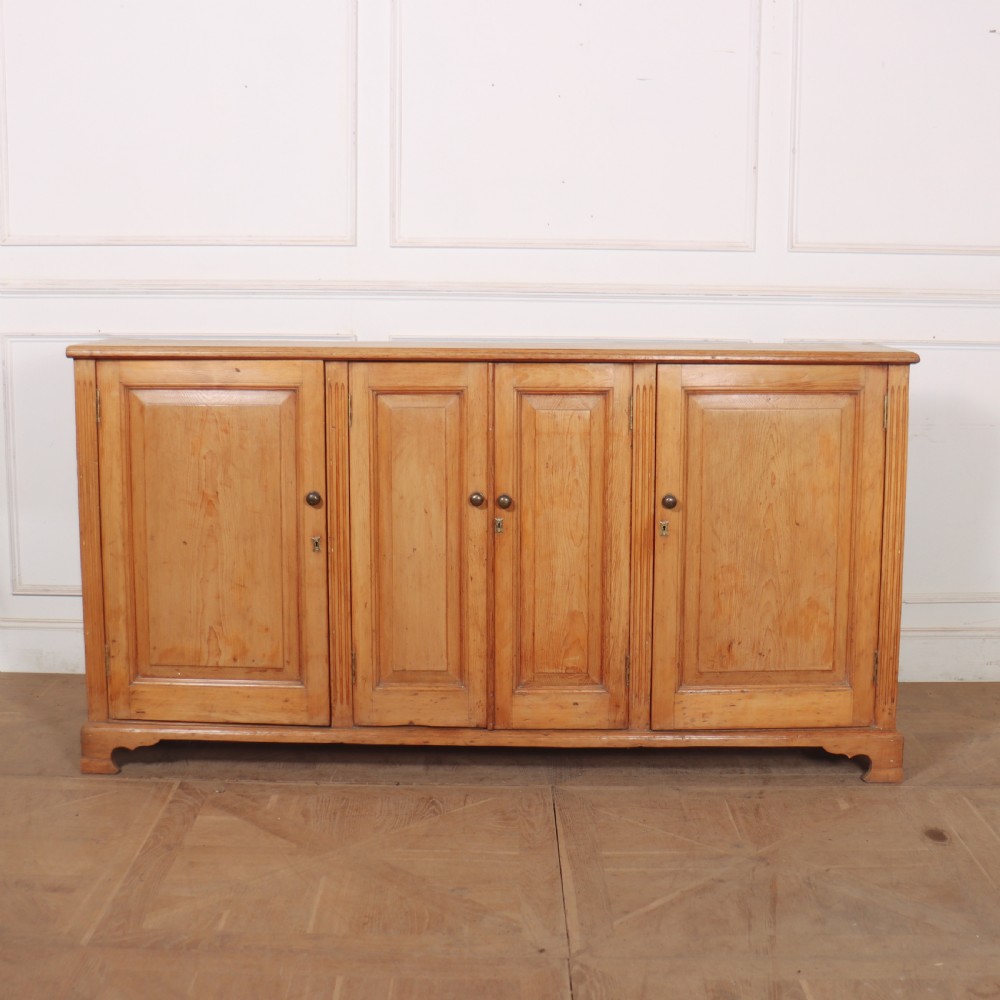 An antique English pine dresser base featuring four fielded cupboard doors with wooden knobs, keyhole escutcheons, fluted vertical details, and bracket feet.