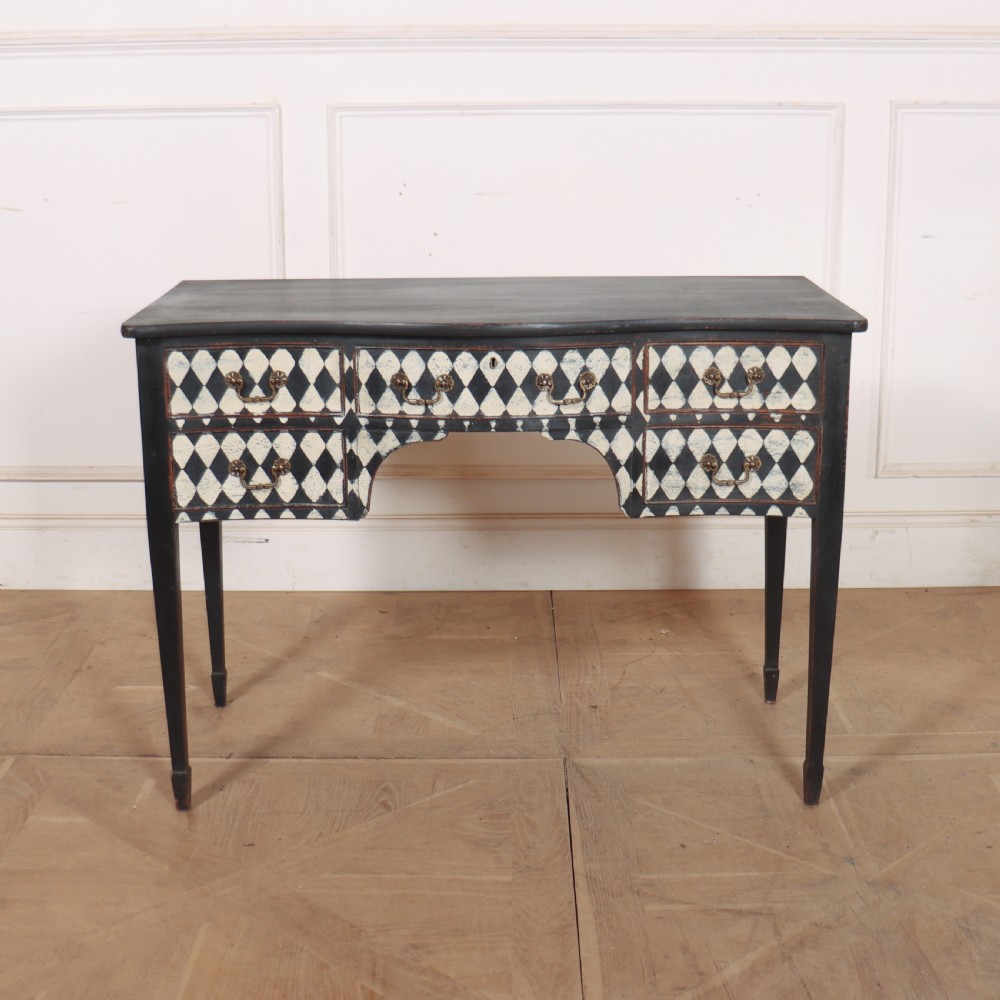 A black painted English serpentine desk featuring a cream and black harlequin pattern on five drawer fronts, brass drop handles, and tapered square legs.