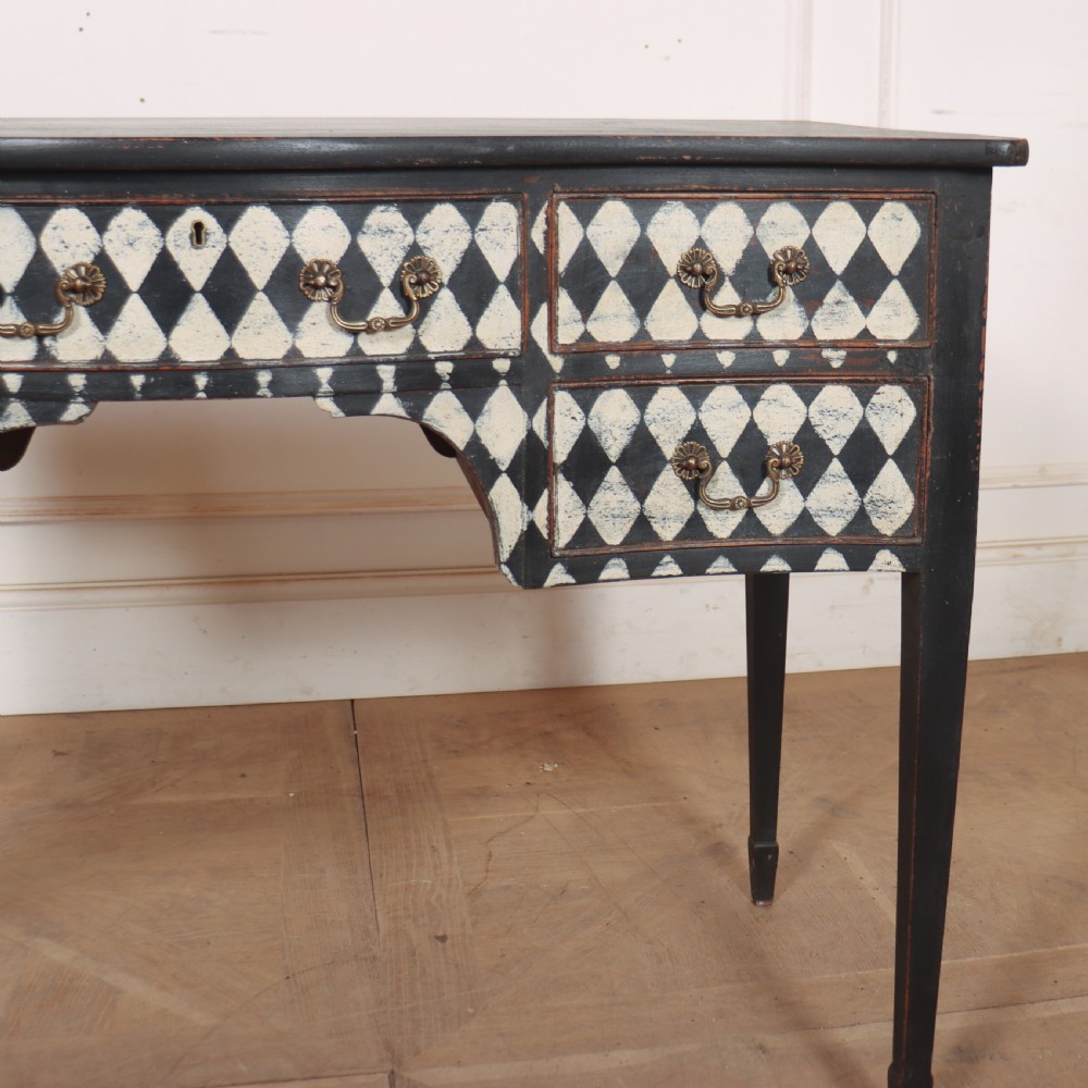 A black painted English serpentine desk featuring a cream and black harlequin pattern on five drawer fronts, brass drop handles, and tapered square legs. View III