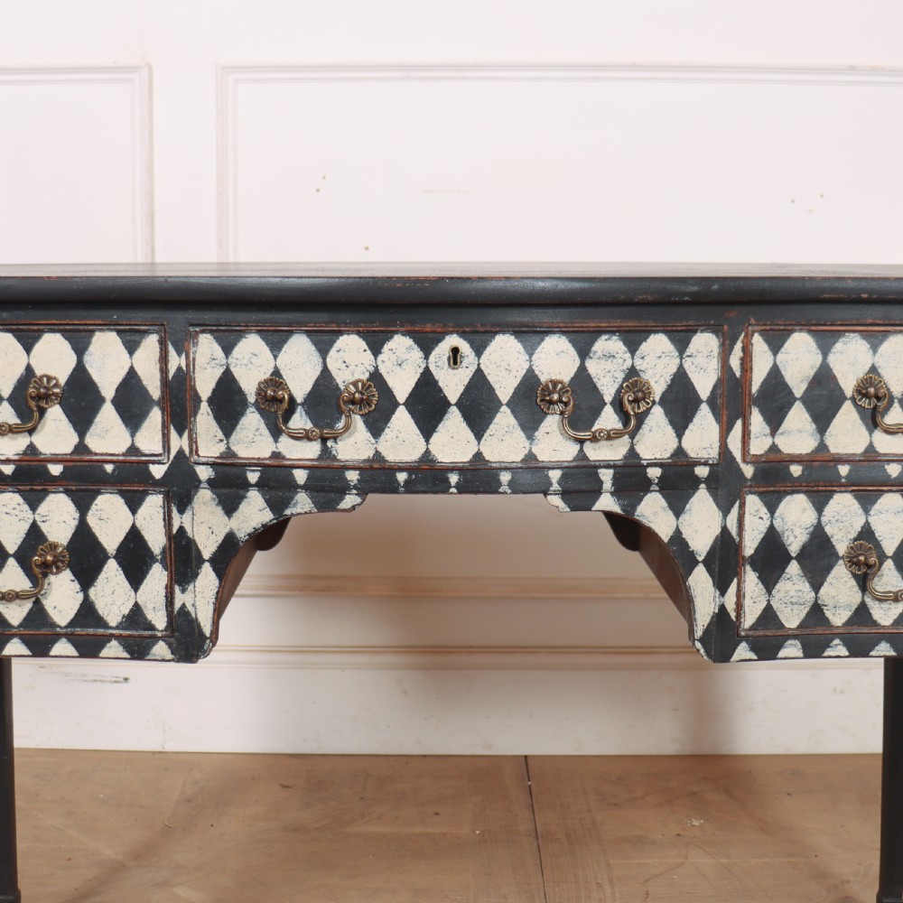 A black painted English serpentine desk featuring a cream and black harlequin pattern on five drawer fronts, brass drop handles, and tapered square legs. View IV