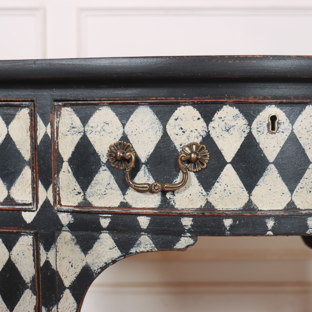 A black painted English serpentine desk featuring a cream and black harlequin pattern on five drawer fronts, brass drop handles, and tapered square legs. View V