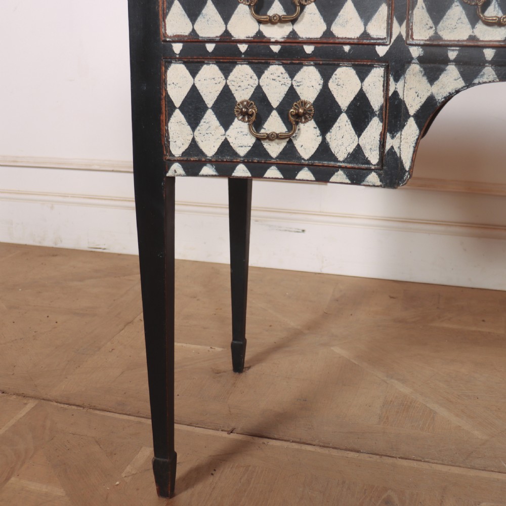 A black painted English serpentine desk featuring a cream and black harlequin pattern on five drawer fronts, brass drop handles, and tapered square legs. View VI