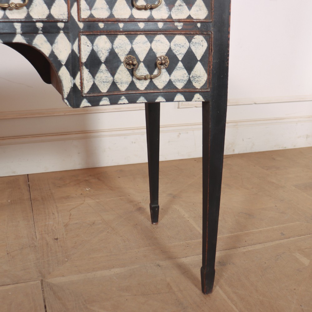 A black painted English serpentine desk featuring a cream and black harlequin pattern on five drawer fronts, brass drop handles, and tapered square legs. View VII