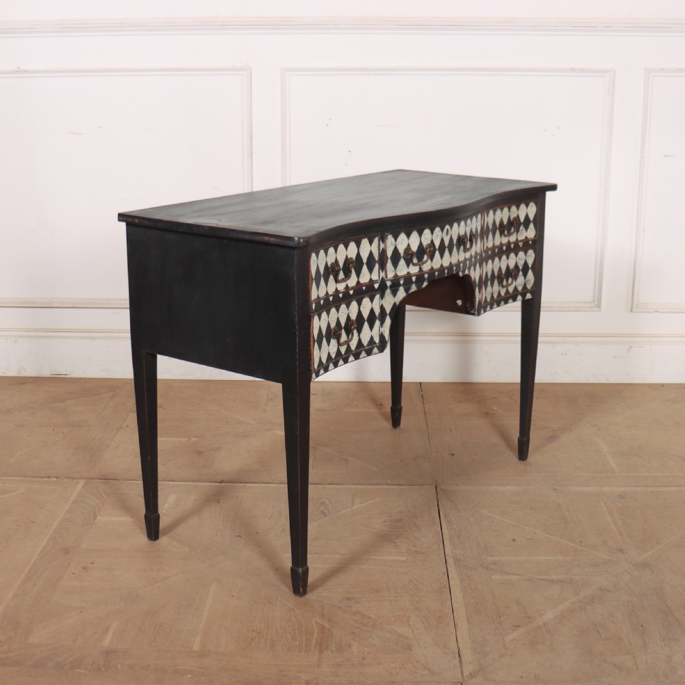 A black painted English serpentine desk featuring a cream and black harlequin pattern on five drawer fronts, brass drop handles, and tapered square legs. View VIII