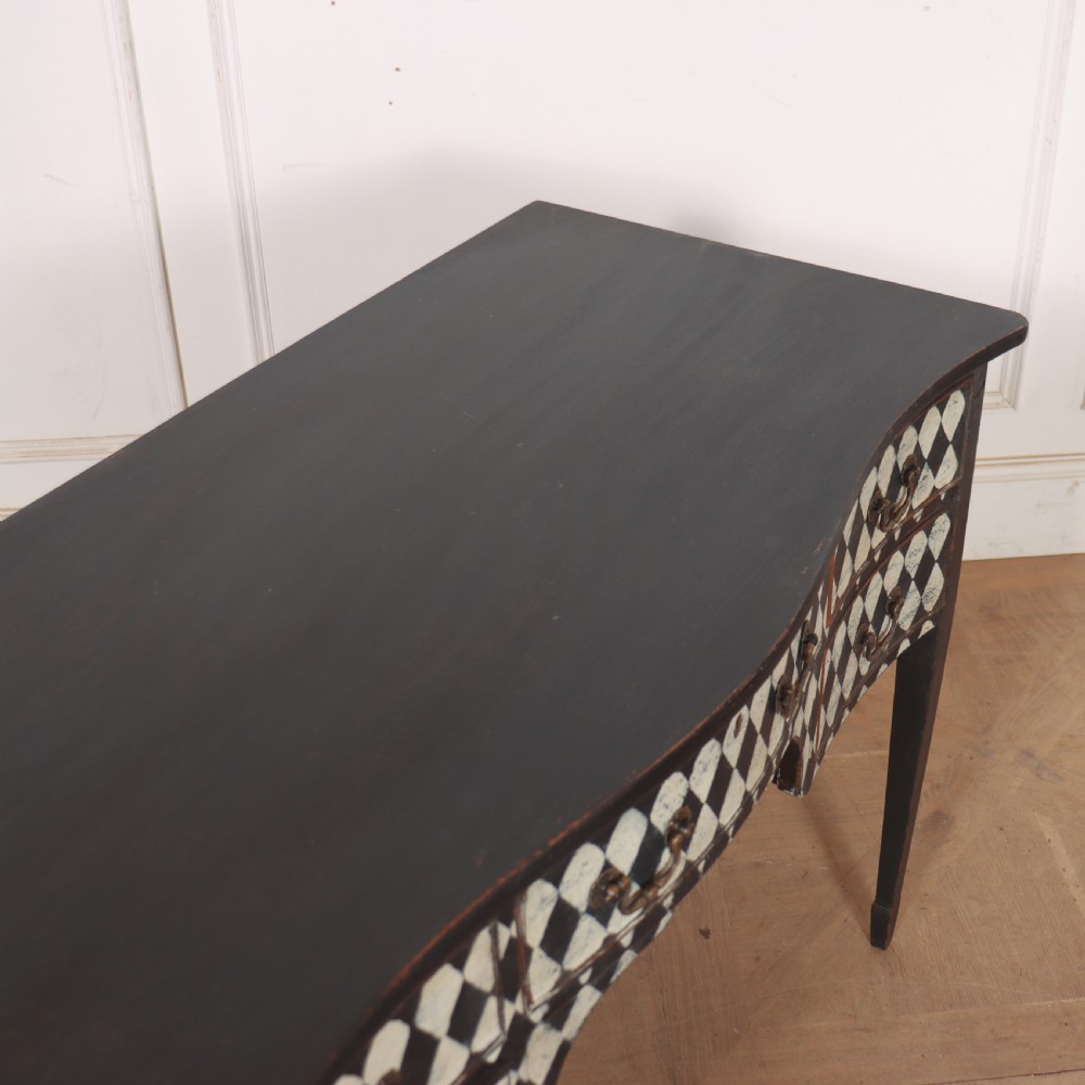 A black painted English serpentine desk featuring a cream and black harlequin pattern on five drawer fronts, brass drop handles, and tapered square legs. View IX