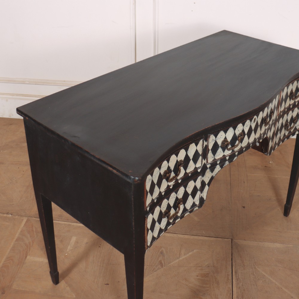 A black painted English serpentine desk featuring a cream and black harlequin pattern on five drawer fronts, brass drop handles, and tapered square legs. View X