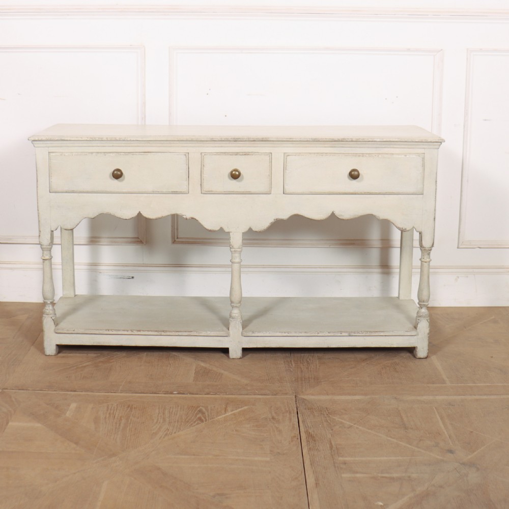 A painted cream timber potboard dresser base featuring three drawers with brass knobs, a scalloped apron, turned legs, and an open lower shelf.