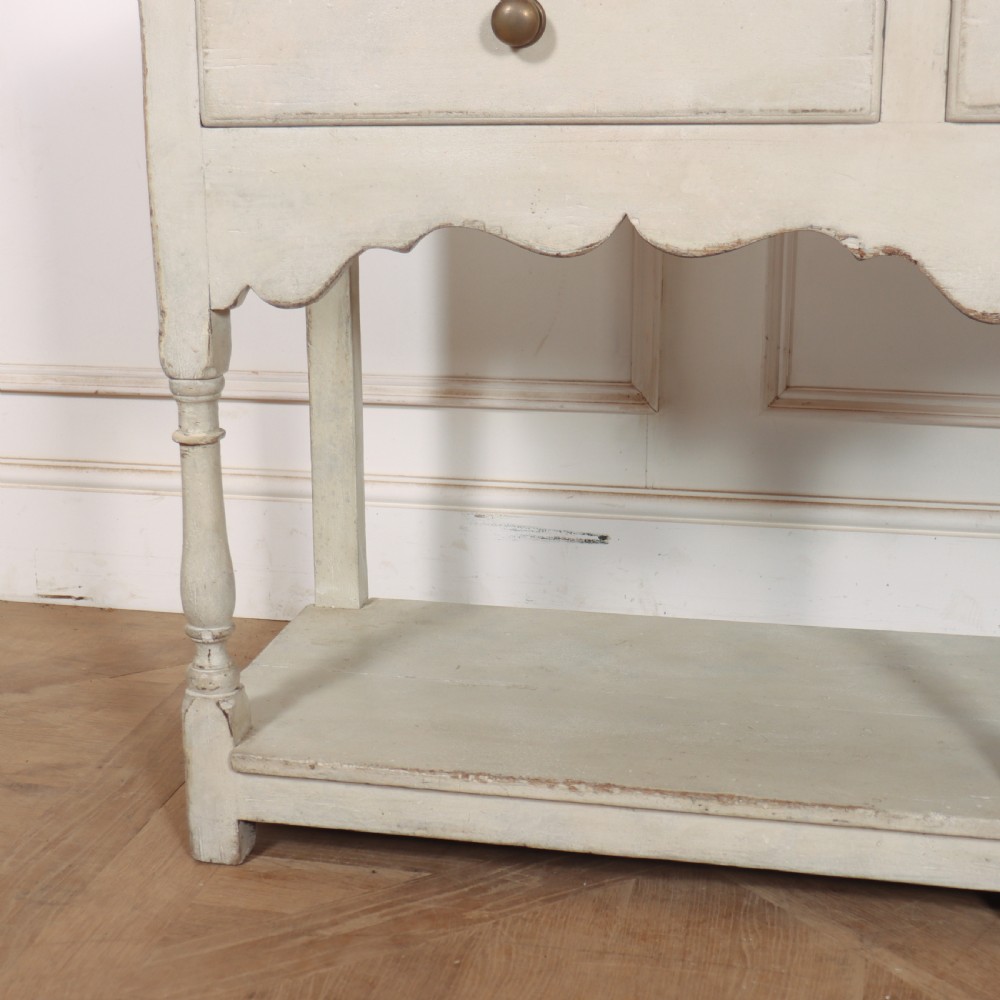 A painted cream timber potboard dresser base featuring three drawers with brass knobs, a scalloped apron, turned legs, and an open lower shelf. View III