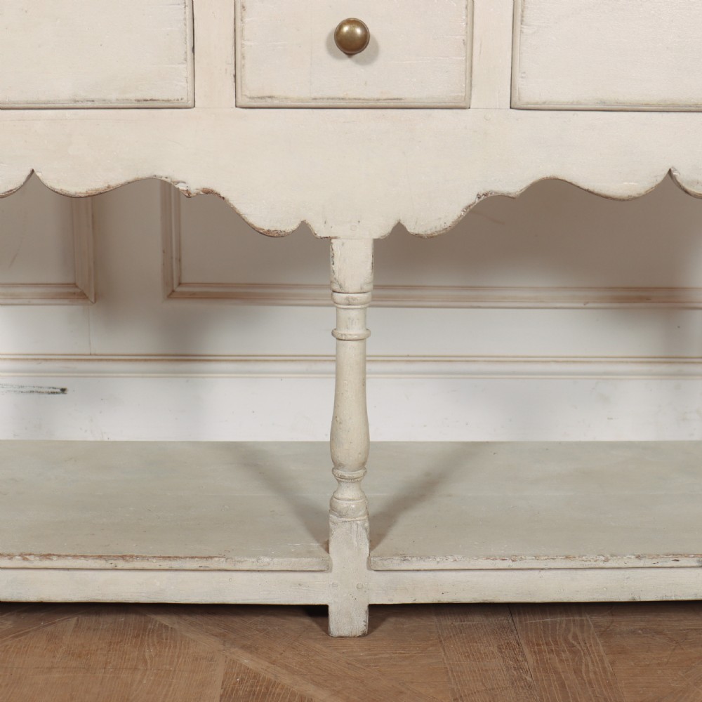 A painted cream timber potboard dresser base featuring three drawers with brass knobs, a scalloped apron, turned legs, and an open lower shelf. View IV