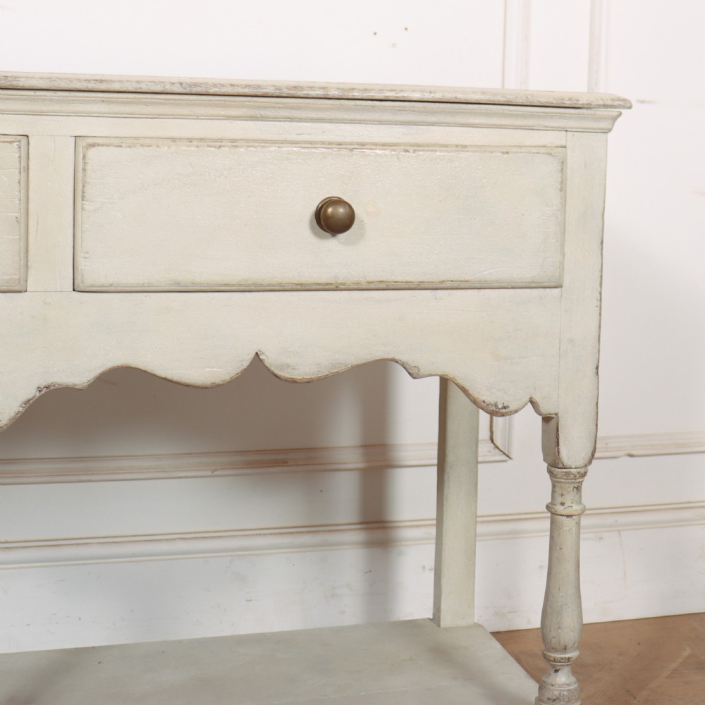 A painted cream timber potboard dresser base featuring three drawers with brass knobs, a scalloped apron, turned legs, and an open lower shelf. View V