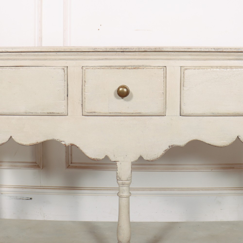 A painted cream timber potboard dresser base featuring three drawers with brass knobs, a scalloped apron, turned legs, and an open lower shelf. View VII