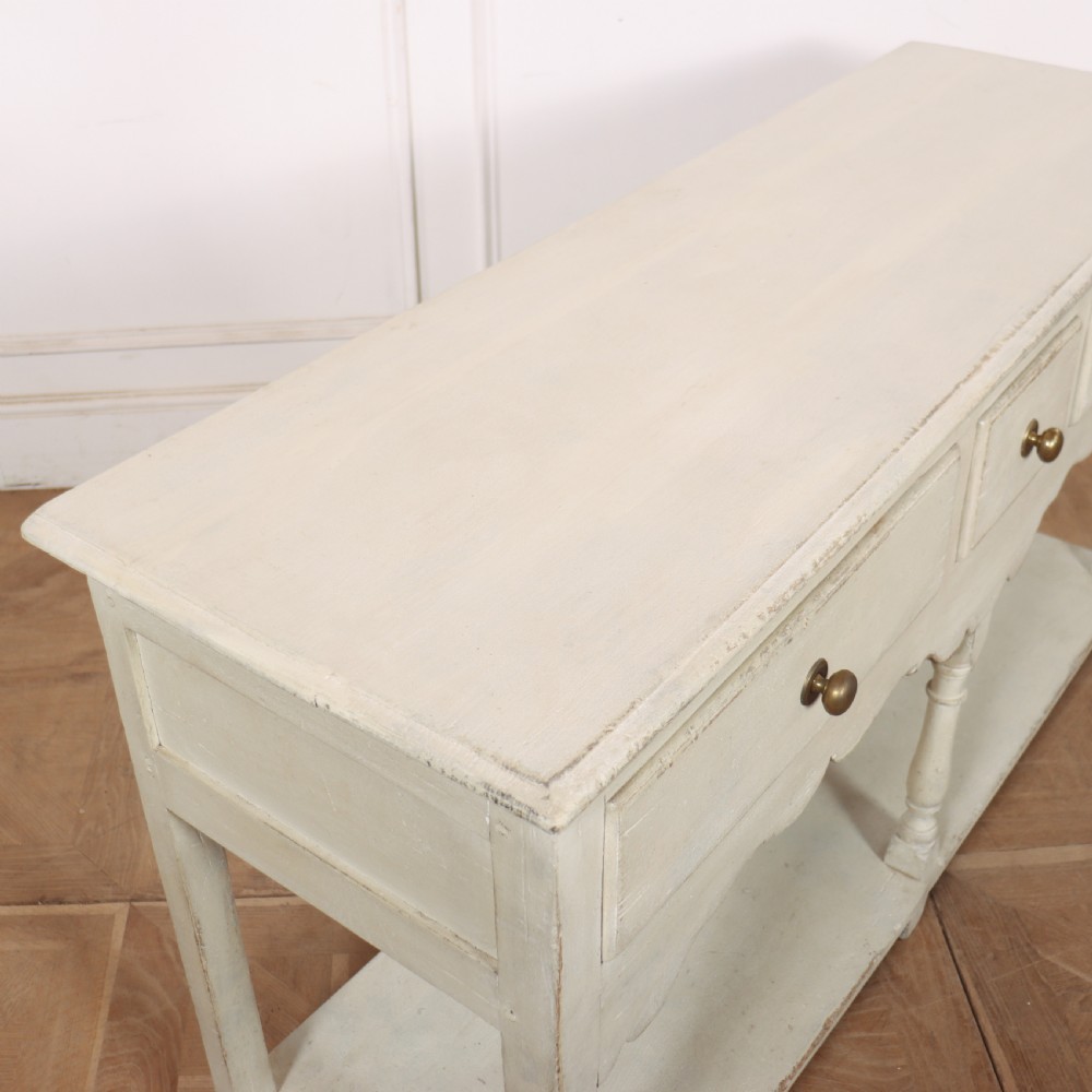 A painted cream timber potboard dresser base featuring three drawers with brass knobs, a scalloped apron, turned legs, and an open lower shelf. View X