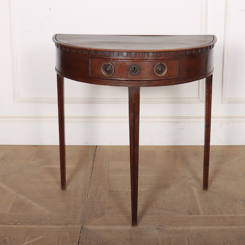 An antique dark oak demi-lune console table featuring three tapered legs, a carved frieze moulding, and a single drawer with brass ring hardware.