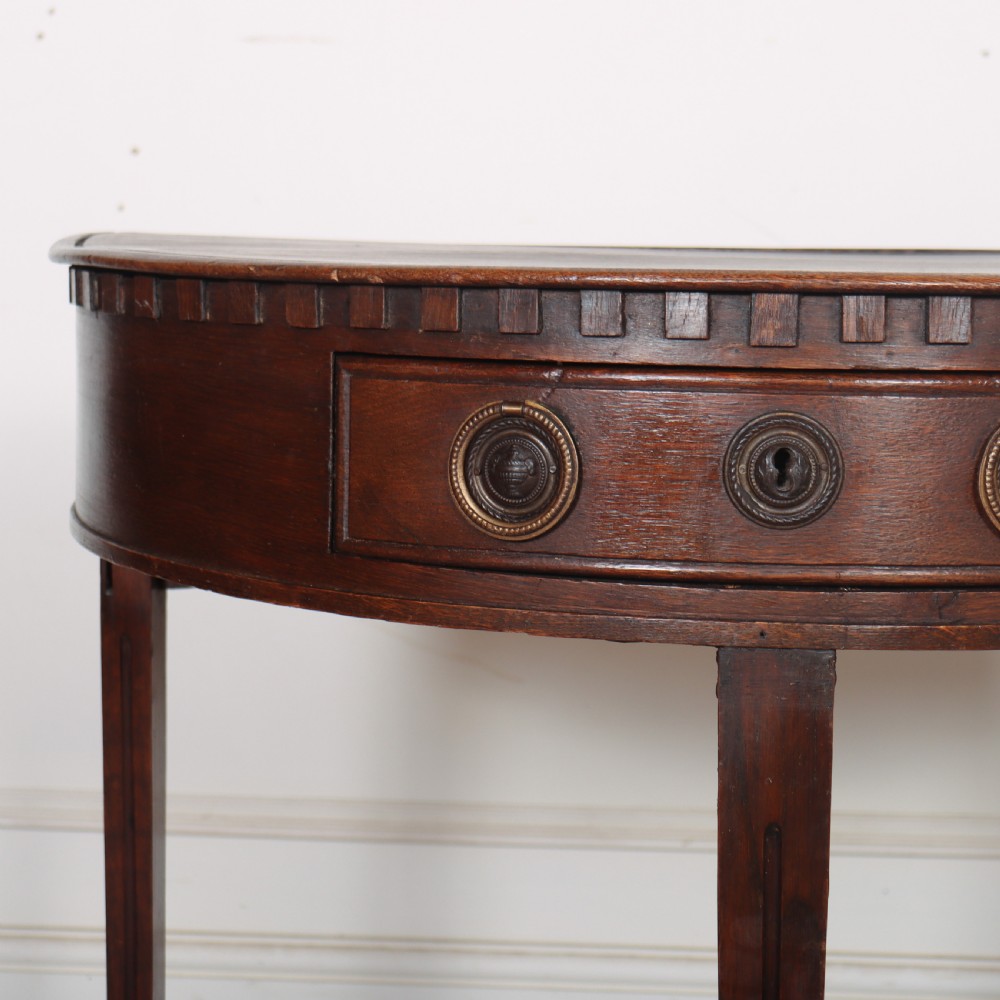 An antique dark oak demi-lune console table featuring three tapered legs, a carved frieze moulding, and a single drawer with brass ring hardware. View II