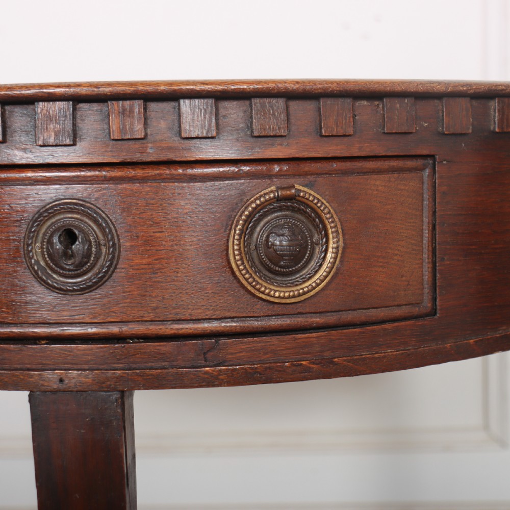 An antique dark oak demi-lune console table featuring three tapered legs, a carved frieze moulding, and a single drawer with brass ring hardware. View III