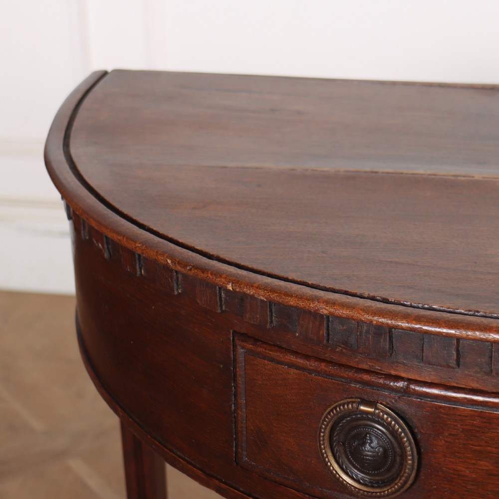 An antique dark oak demi-lune console table featuring three tapered legs, a carved frieze moulding, and a single drawer with brass ring hardware. View IV