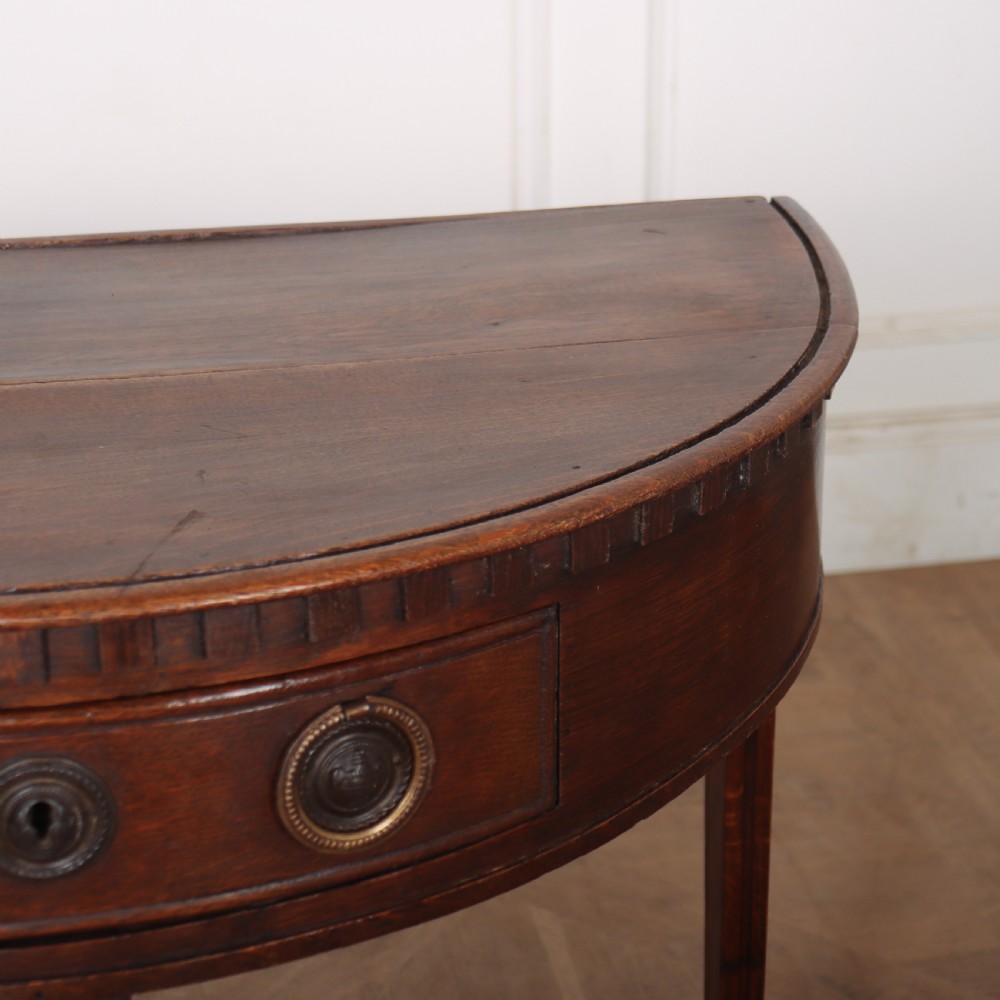An antique dark oak demi-lune console table featuring three tapered legs, a carved frieze moulding, and a single drawer with brass ring hardware. View V