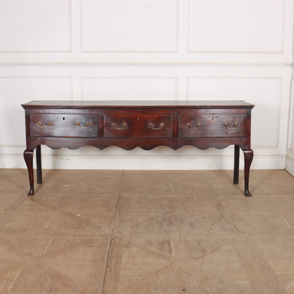 An 18th-century English oak dresser base featuring three drawers with brass handles, a scalloped apron, and cabriole legs.