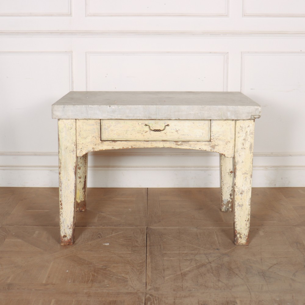 An antique pastry table featuring a distressed cream painted wooden frame, a single central drawer with a brass handle, and a thick grey metal top.