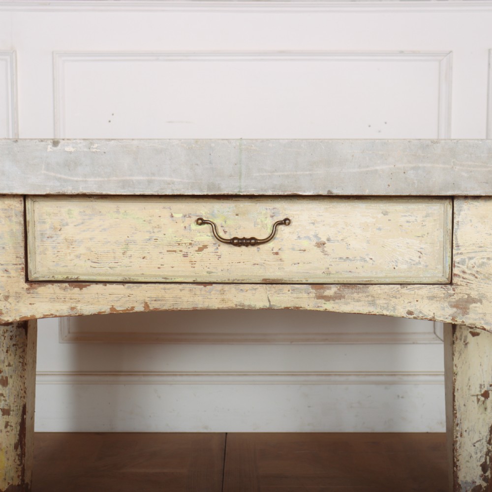 An antique pastry table featuring a distressed cream painted wooden frame, a single central drawer with a brass handle, and a thick grey metal top. View V
