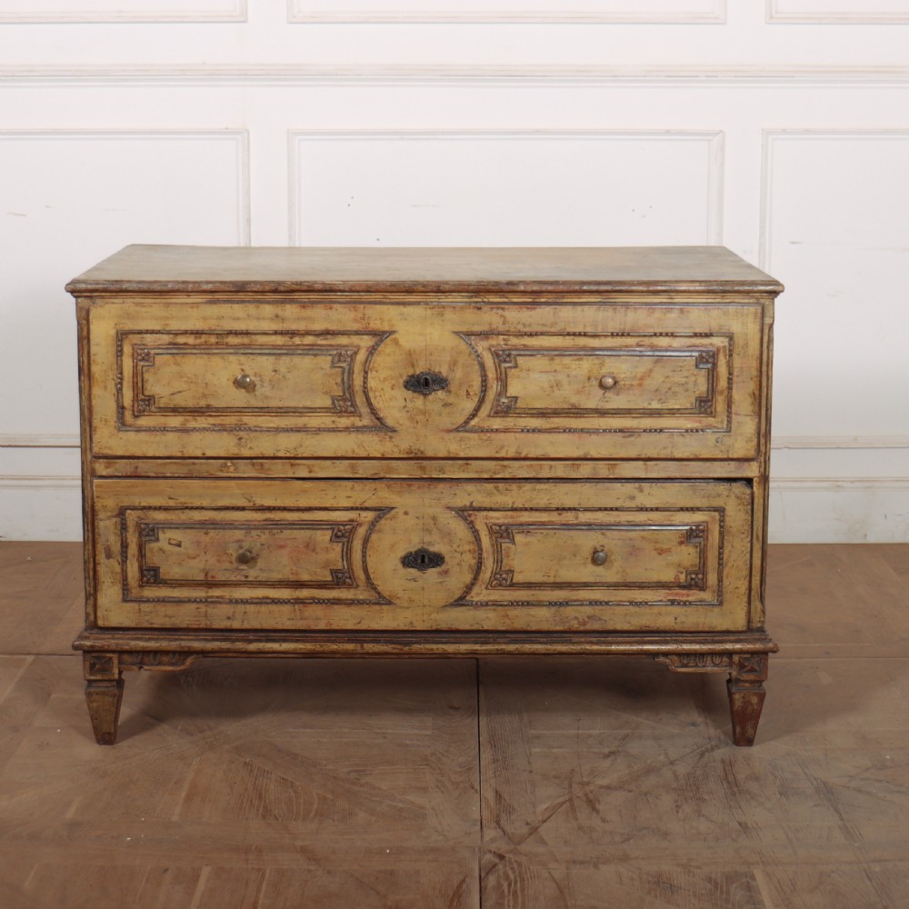 An antique two-drawer Italian commode with a muted yellow painted distressed finish, geometric panel mouldings, central brass escutcheons, and square tapered legs.