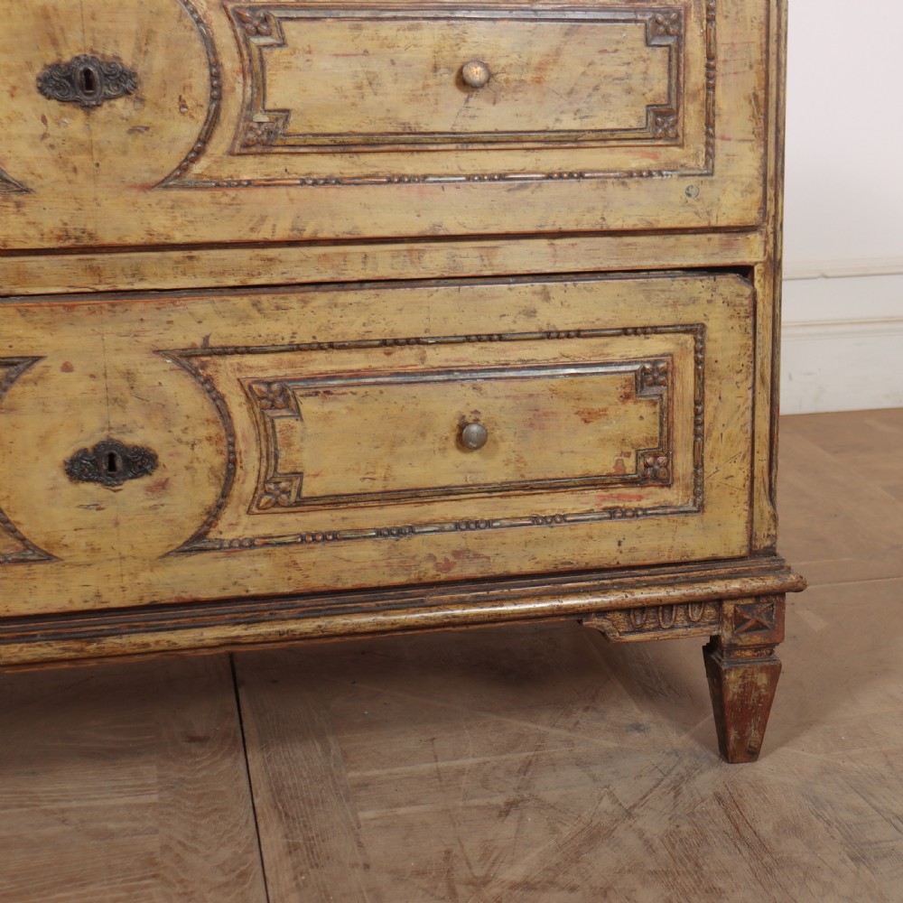 An antique two-drawer Italian commode with a muted yellow painted distressed finish, geometric panel mouldings, central brass escutcheons, and square tapered legs. View VI