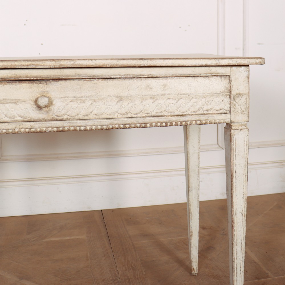 An antique off-white painted Swedish side table featuring tapered fluted legs, carved rope detail along the drawer frieze, and a single central drawer pull. View III