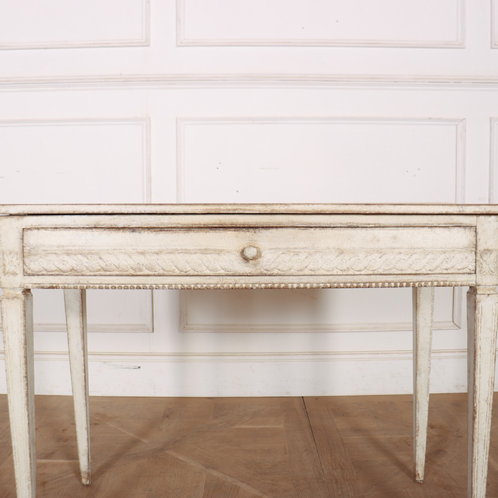 An antique off-white painted Swedish side table featuring tapered fluted legs, carved rope detail along the drawer frieze, and a single central drawer pull. View VII