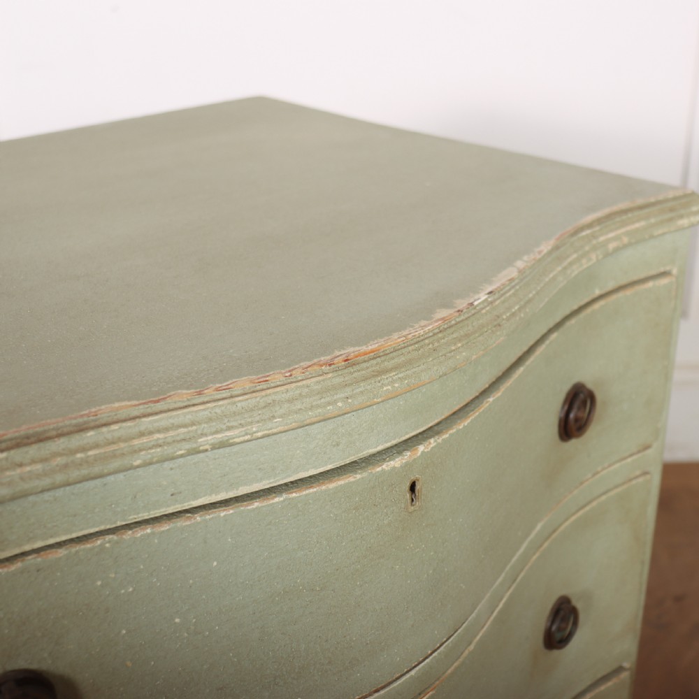 A Swedish serpentine painted commode featuring three drawers with brass pulls and a shaped apron in a muted green finish. View IX