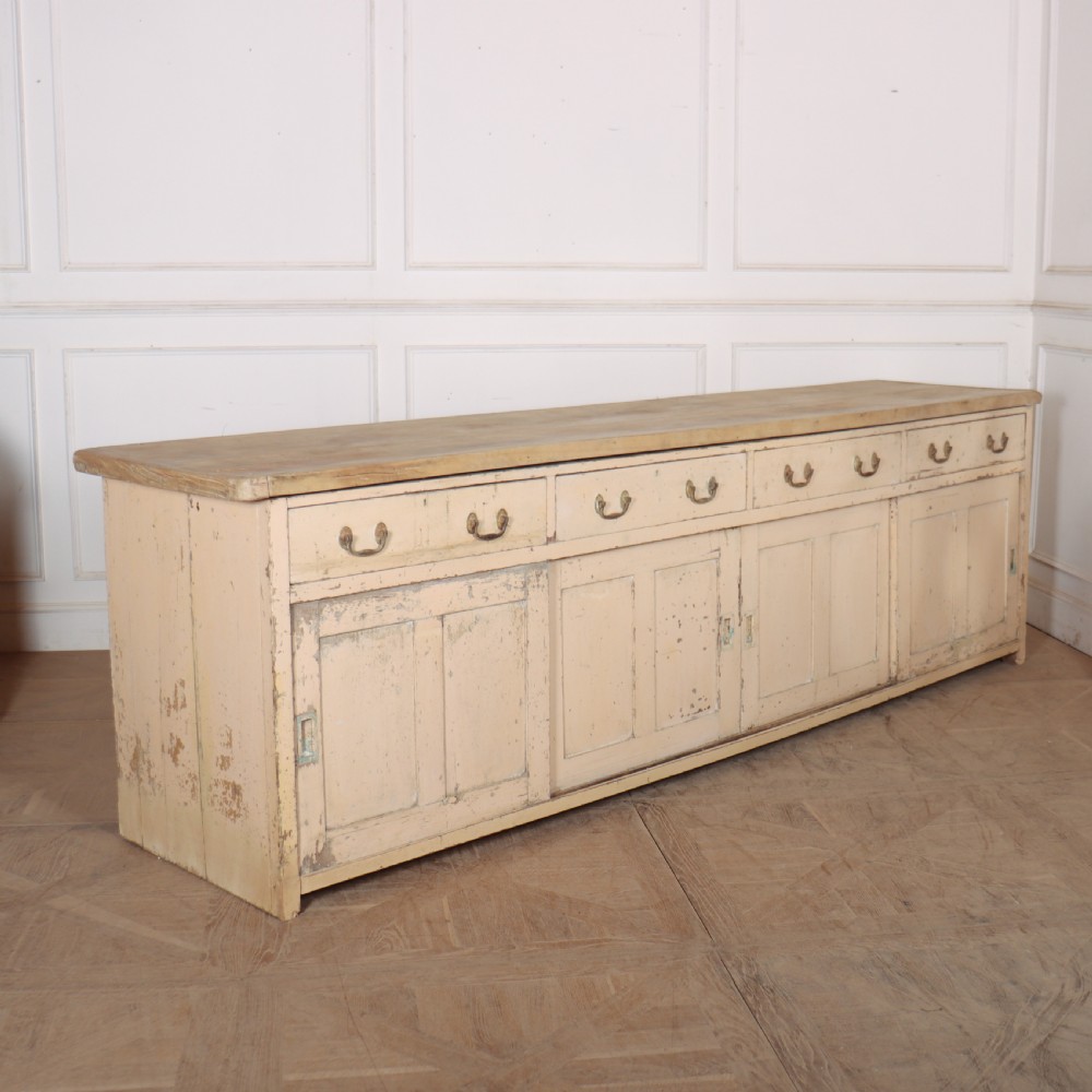 A long antique painted pine dresser base featuring four upper drawers with brass handles above panelled cupboard doors and a natural timber top.