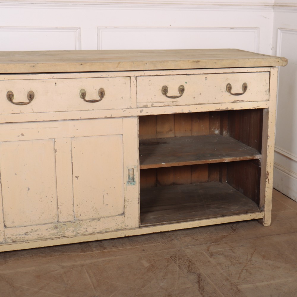 A long antique painted pine dresser base featuring four upper drawers with brass handles above panelled cupboard doors and a natural timber top. View X