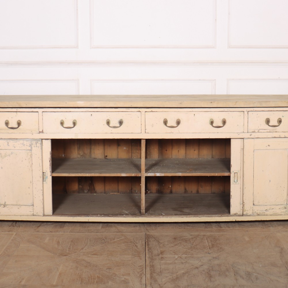 A long antique painted pine dresser base featuring four upper drawers with brass handles above panelled cupboard doors and a natural timber top. View XI