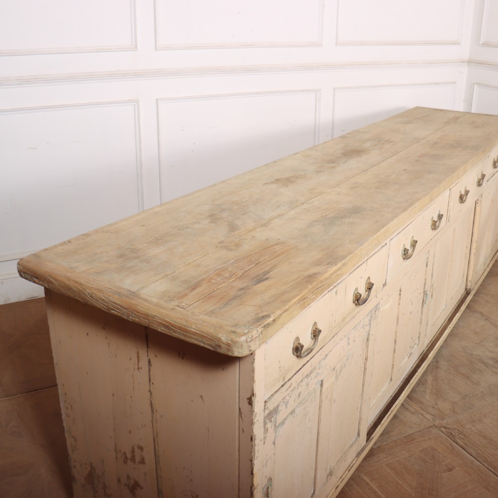 A long antique painted pine dresser base featuring four upper drawers with brass handles above panelled cupboard doors and a natural timber top. View XII