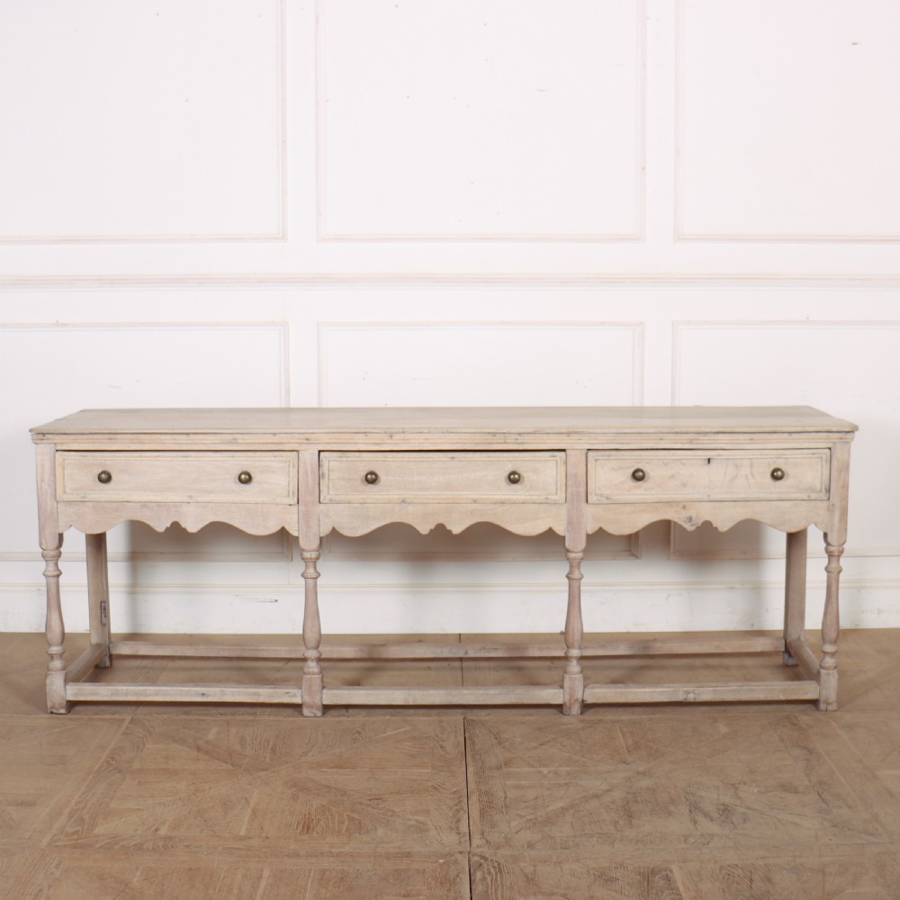 An English bleached oak dresser base featuring three drawers with brass knob handles, a scalloped apron, and turned legs connected by a stretcher base.
