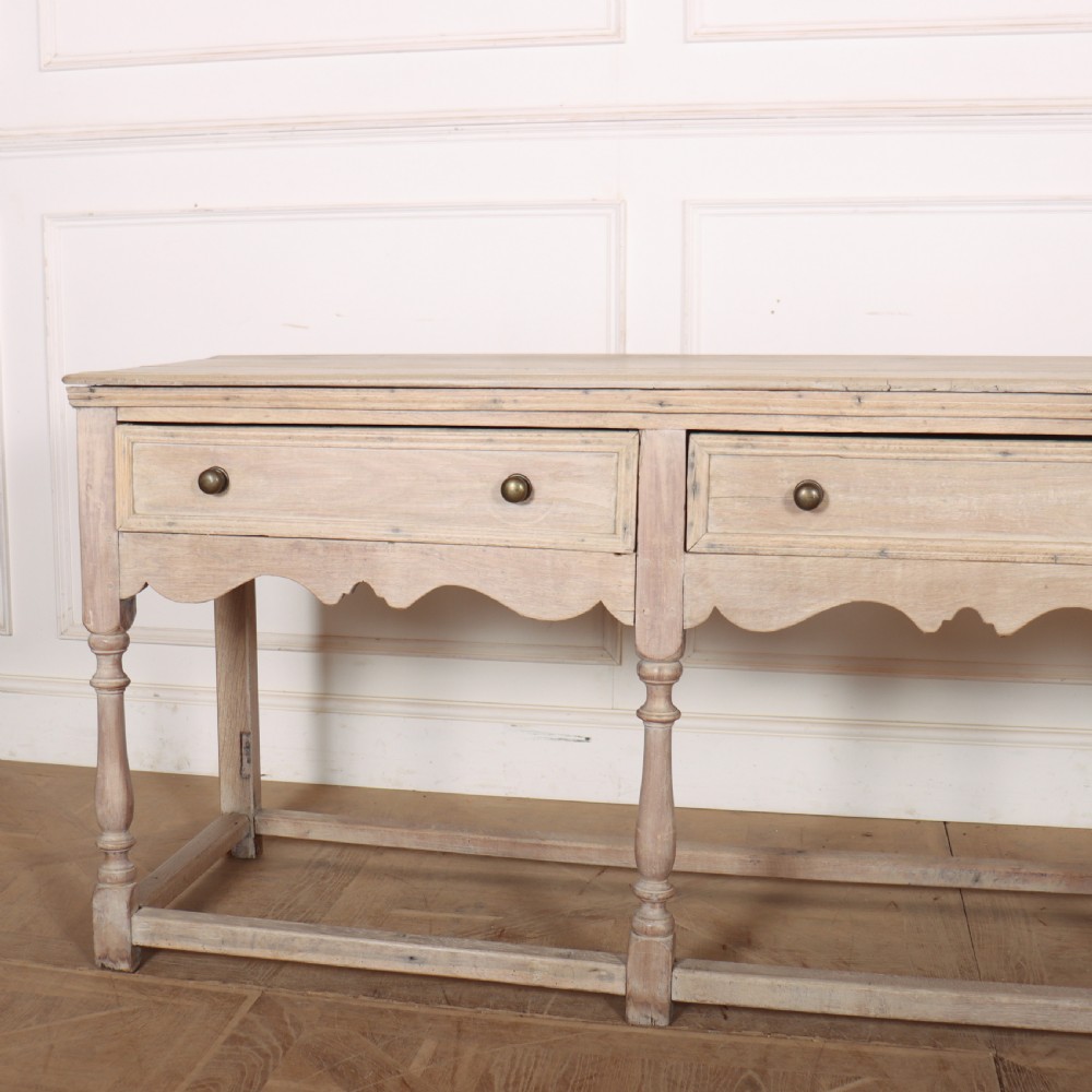 An English bleached oak dresser base featuring three drawers with brass knob handles, a scalloped apron, and turned legs connected by a stretcher base. View II