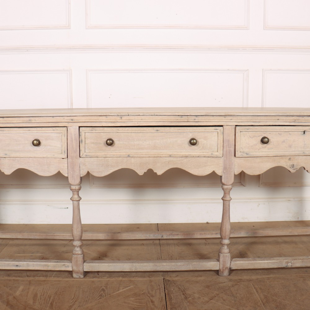 An English bleached oak dresser base featuring three drawers with brass knob handles, a scalloped apron, and turned legs connected by a stretcher base. View III