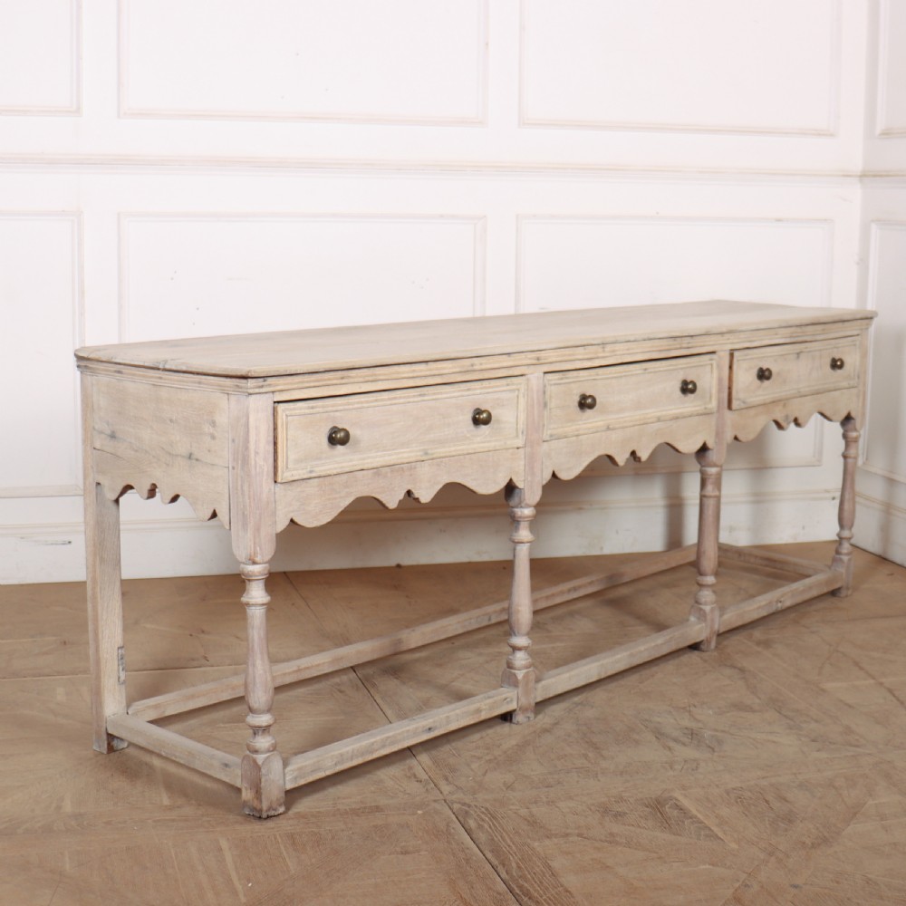 An English bleached oak dresser base featuring three drawers with brass knob handles, a scalloped apron, and turned legs connected by a stretcher base. View VII