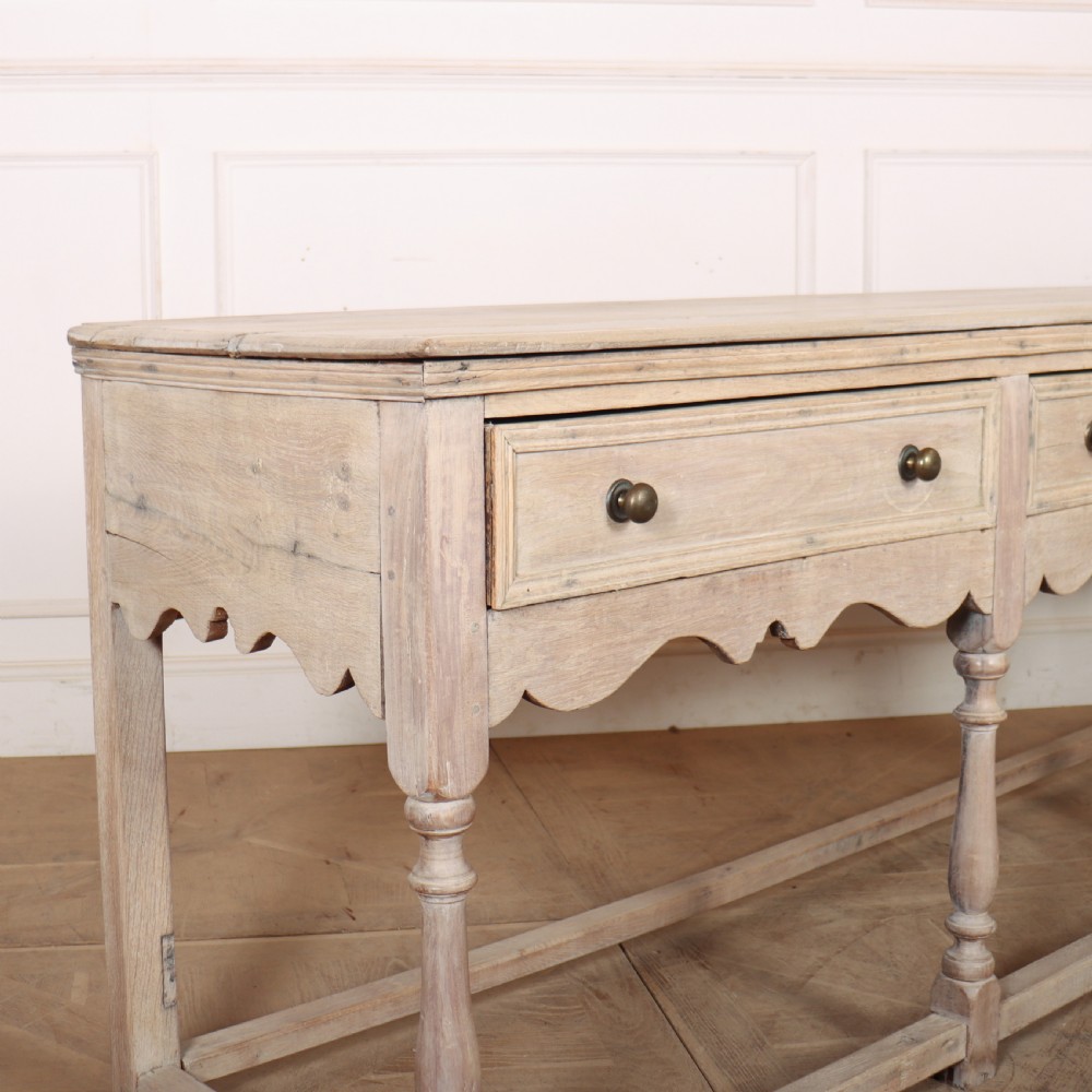 An English bleached oak dresser base featuring three drawers with brass knob handles, a scalloped apron, and turned legs connected by a stretcher base. View IX