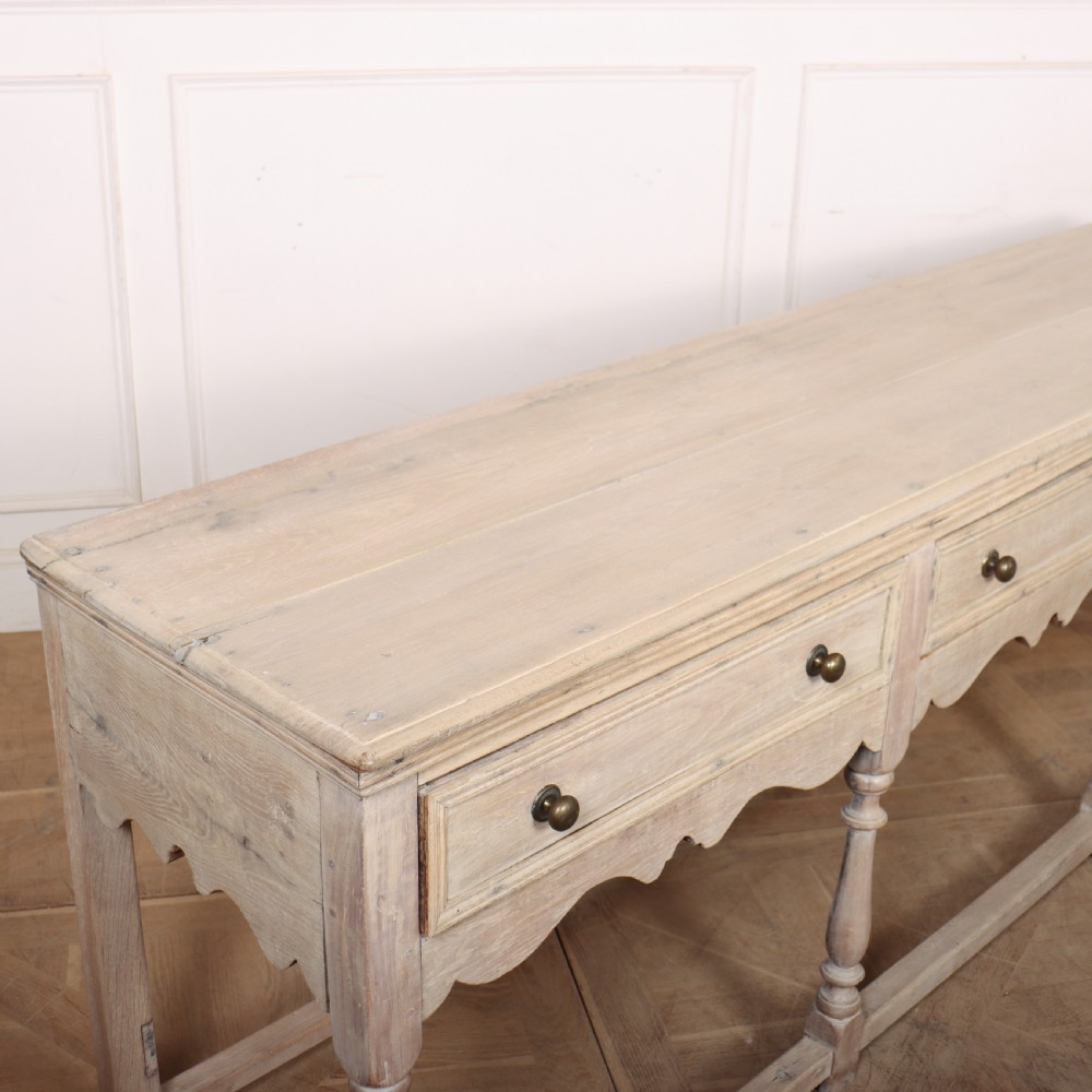 An English bleached oak dresser base featuring three drawers with brass knob handles, a scalloped apron, and turned legs connected by a stretcher base. View X