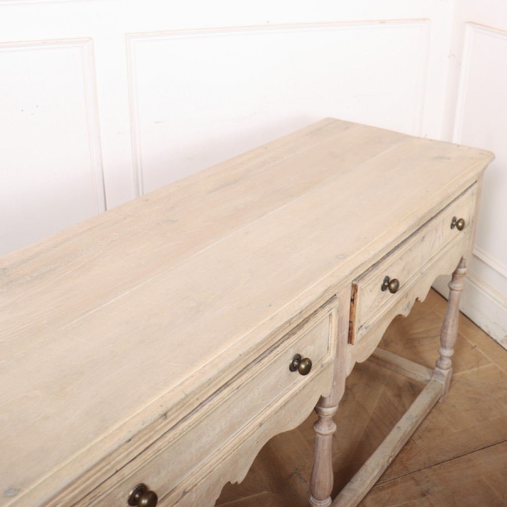 An English bleached oak dresser base featuring three drawers with brass knob handles, a scalloped apron, and turned legs connected by a stretcher base. View XI