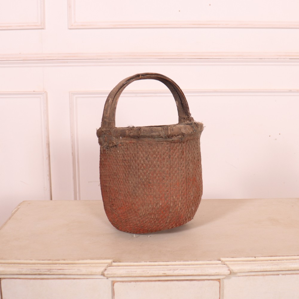An antique English woven basket featuring a terracotta red painted finish, a structured rim, and a sturdy upright handle.