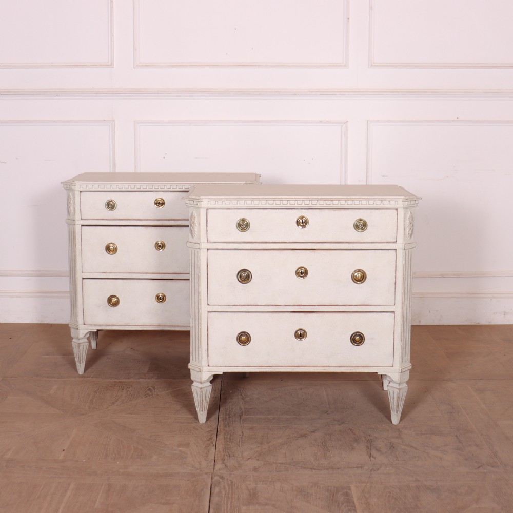 A pair of Swedish Gustavian style off-white painted commodes featuring three drawers each, brass hardware, fluted sides, and square tapered feet.