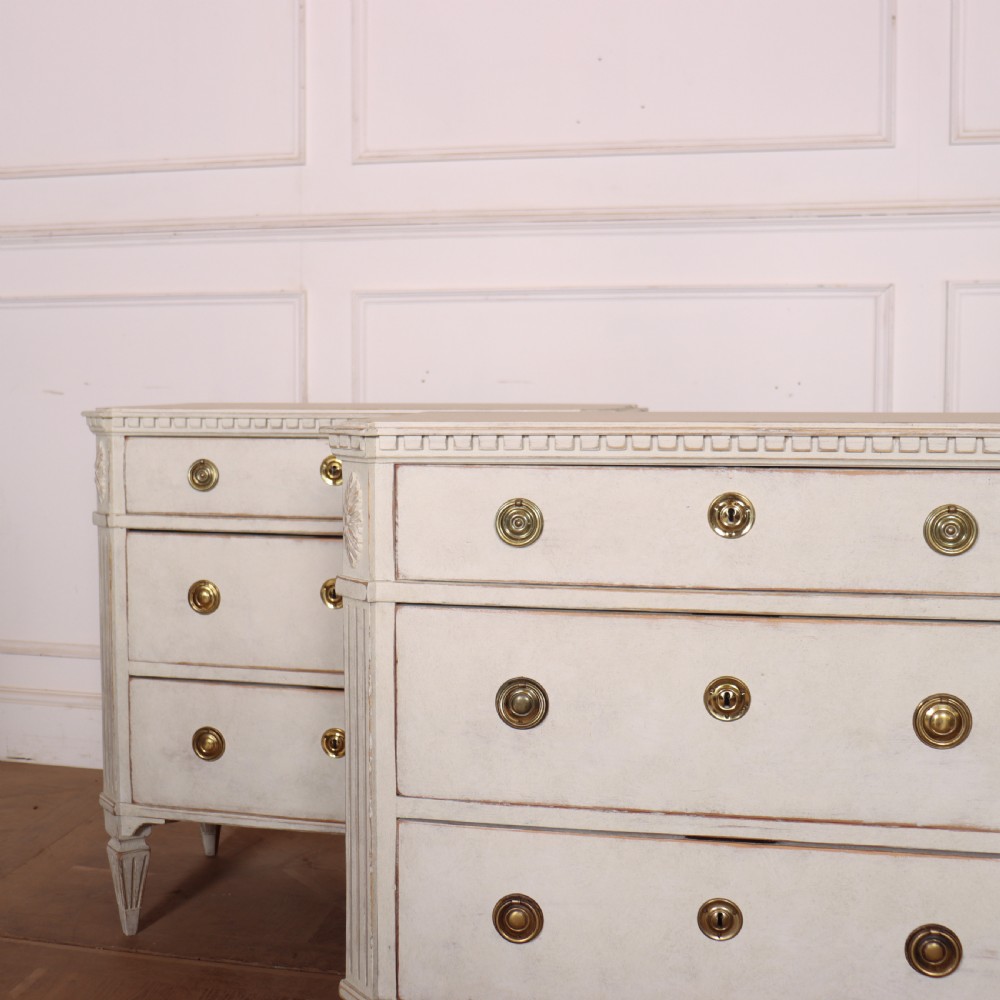 A pair of Swedish Gustavian style off-white painted commodes featuring three drawers each, brass hardware, fluted sides, and square tapered feet. View II