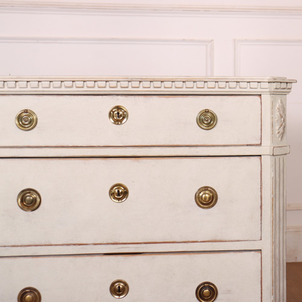 A pair of Swedish Gustavian style off-white painted commodes featuring three drawers each, brass hardware, fluted sides, and square tapered feet. View IV