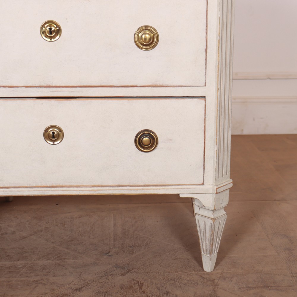 A pair of Swedish Gustavian style off-white painted commodes featuring three drawers each, brass hardware, fluted sides, and square tapered feet. View V